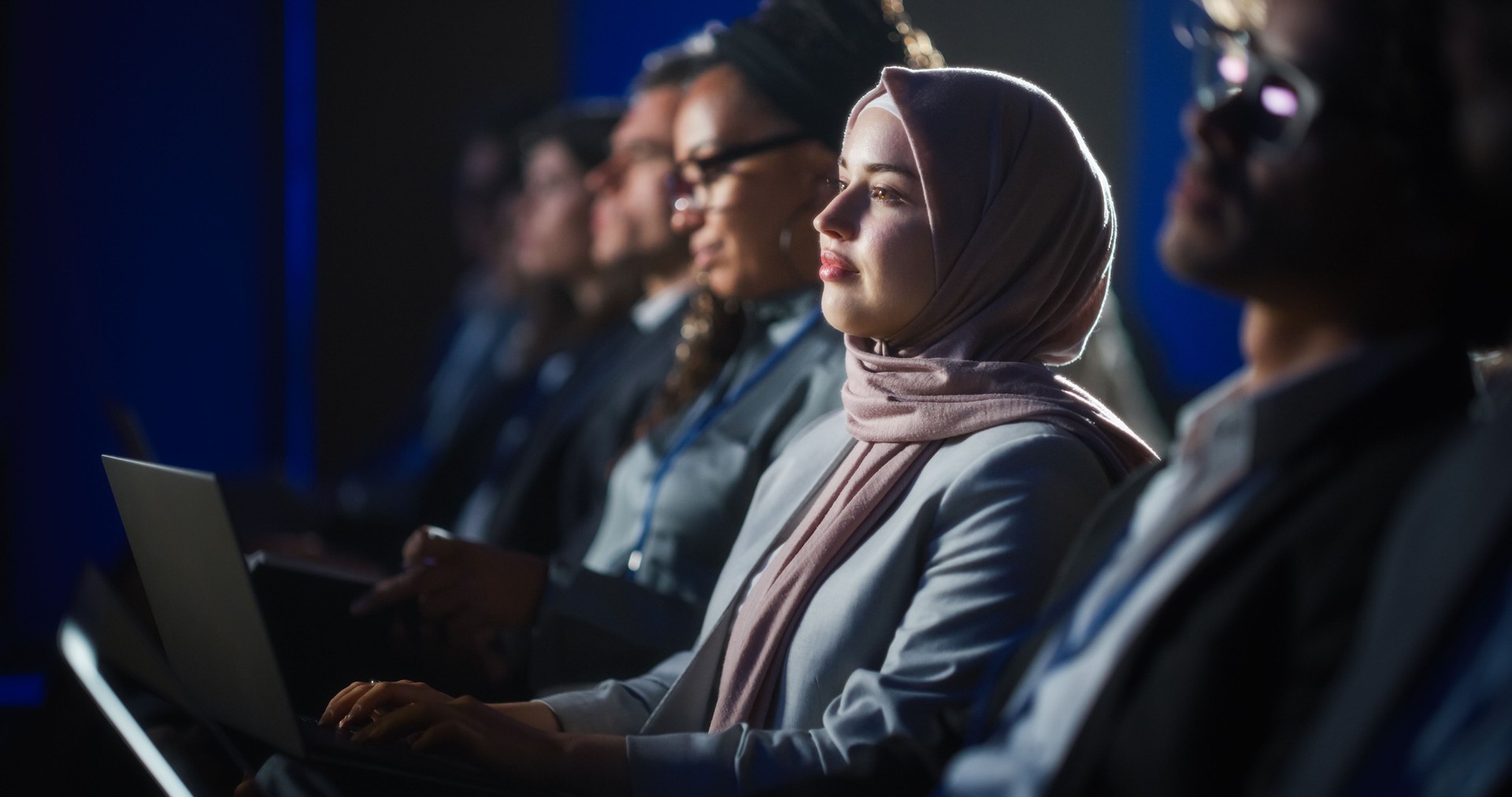 Arab Female Sitting in a Dark Crowded Auditorium at a Human Rights Conference. Young Muslim Woman Using Laptop Computer. Activist in Hijab Listening to Inspiring Speech About Global Initiative.